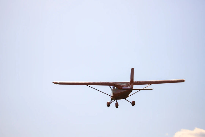 A small high-wing airplane flying against a pale sky, seen from head on