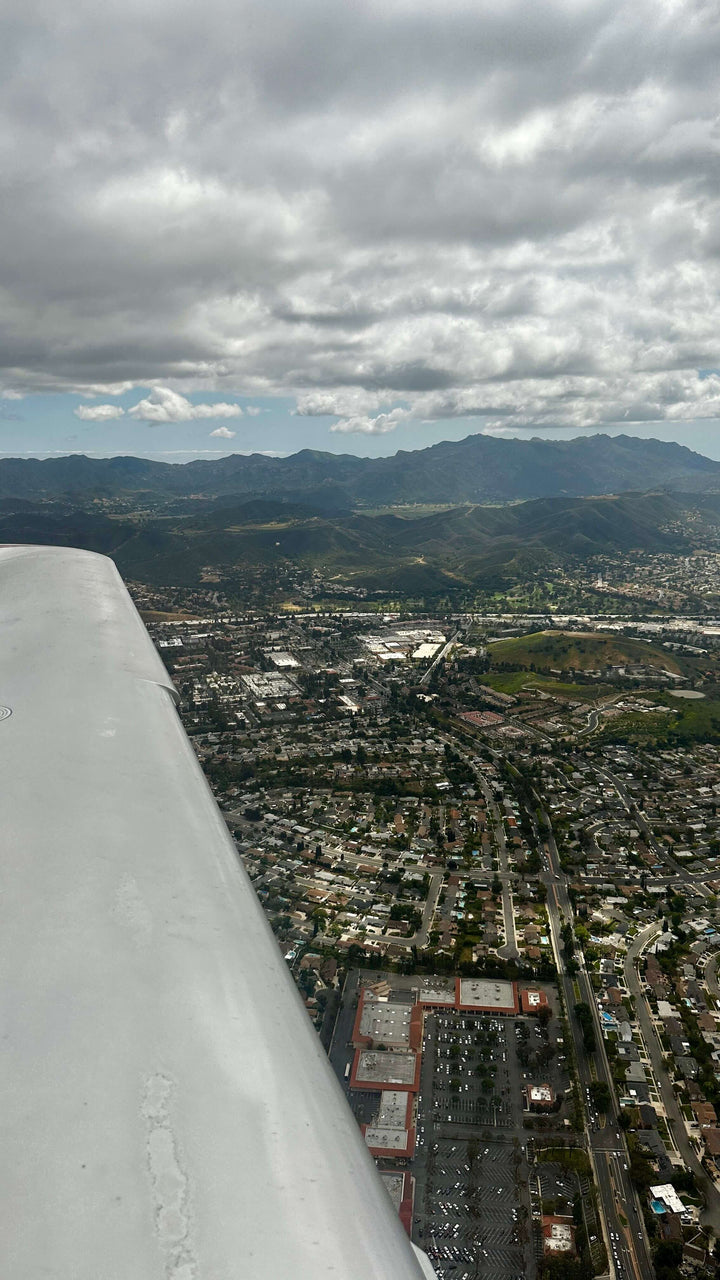 The wing of a small airplane over the San Fernando Valley on the way back to Van Nuys, with the city grid below and mountains under the clouds on the horizon