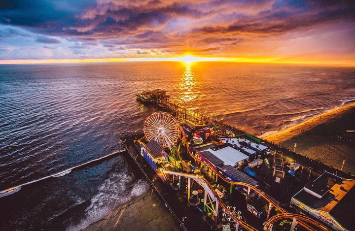 Santa Monica Pier and its Ferris wheel from the air at sunset, with the sun going down over the Pacific Ocean and waves breaking along the beach