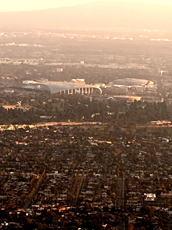 SoFi Stadium in Inglewood at golden hour, seen from a small airplane passing just north of it, with the neighborhoods of South Los Angeles in the foreground