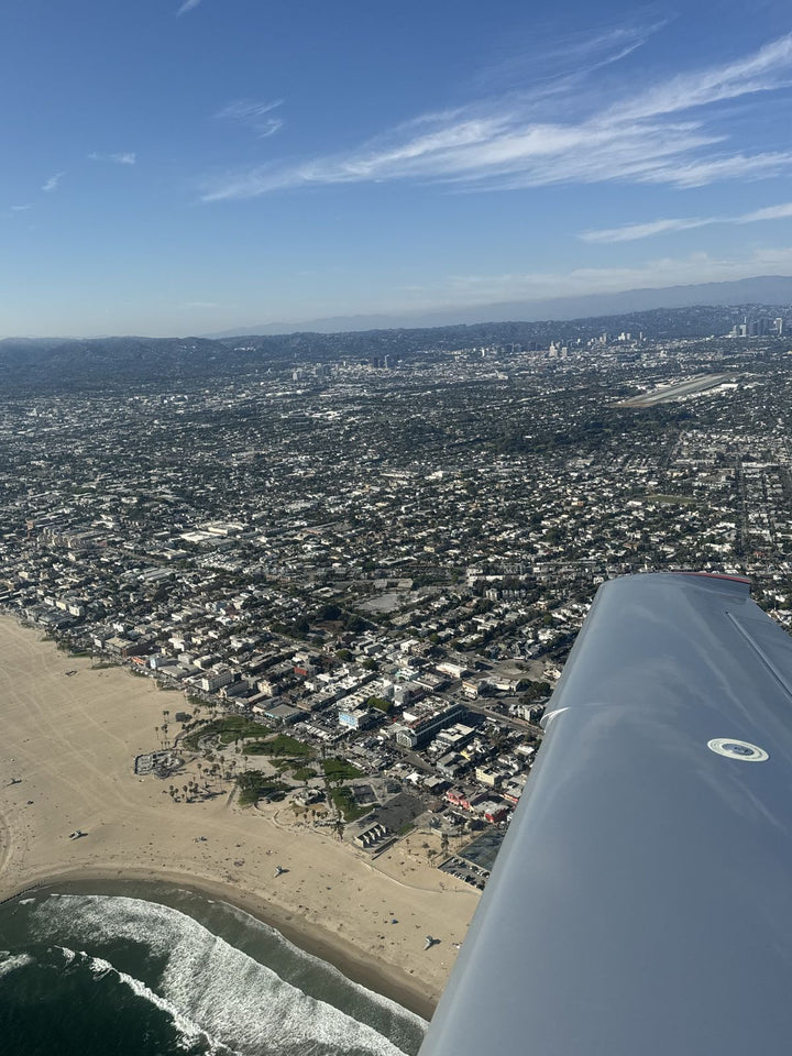 Venice Beach seen over the wing of a small airplane, with the wide sand, the surf and the boardwalk, and the Westside stretching inland to the hills